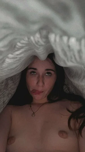 Woman with dark hair under white blanket, bare chest, wearing necklace, looking at camera.