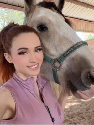 Red-haired woman in purple top poses with horse in stable.