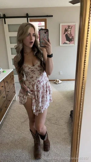 Woman with long wavy hair in floral dress and cowboy boots, taking mirror selfie in bedroom.