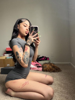 Woman with tattoos in gray dress kneeling on carpet, taking mirror selfie in room.