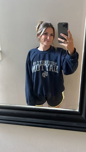Woman in navy sweatshirt and black shorts taking mirror selfie indoors.
