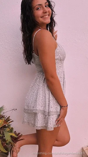Woman in floral dress posing by pink wall with plants, smiling.