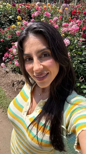Woman with long dark hair in striped shirt, smiling in front of rose garden.