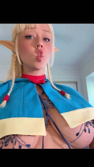 Woman with blonde braids and elf ears in blue costume, posing indoors with lace top.