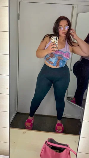 Woman in colorful crop top and dark leggings takes mirror selfie in bedroom.