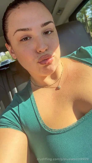 Woman in green top taking selfie in car, hair pulled back, wearing necklace.