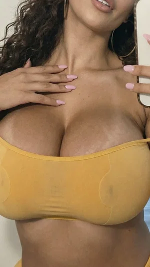 Woman with curly hair in yellow top, holding phone for mirror selfie, indoor setting.