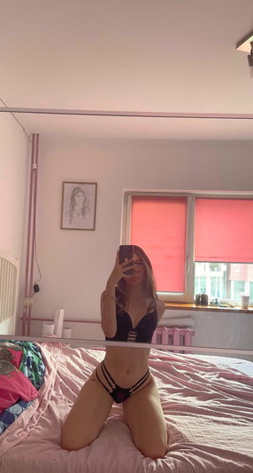 Woman in black lingerie kneeling on bed taking mirror selfie in bedroom.