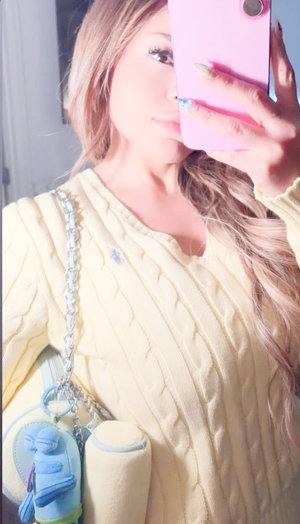 Woman with long blonde hair in yellow sweater holding pink phone, carrying small bag with blue
