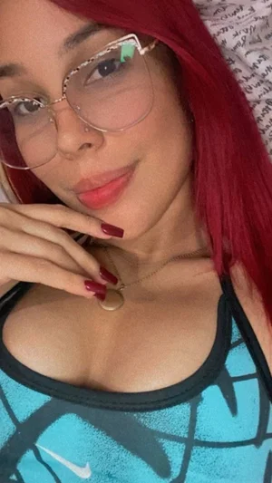 Woman with red hair and glasses wearing a blue tank top, posing indoors.