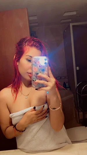 Woman with red hair wrapped in towel, holding phone for mirror selfie in bathroom.