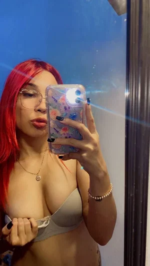 Woman with red hair and glasses in a gray bra taking mirror selfie indoors.