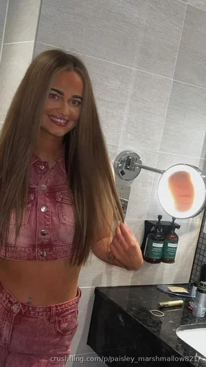 Woman with long brown hair in pink denim crop top and matching pants, standing in bathroom.