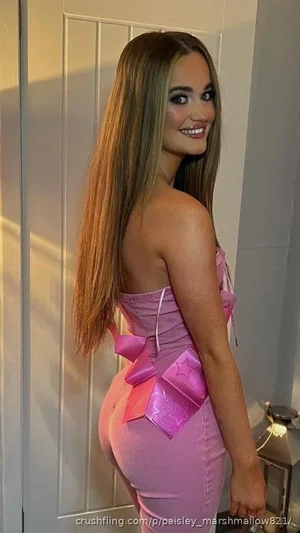 Woman in pink outfit with bow detail, standing indoors near door, smiling at camera.