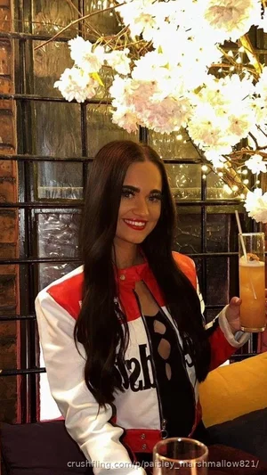 Woman with long dark hair in red and white jacket holding drink, seated indoors near floral lights.