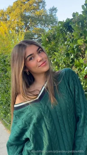 Woman with long hair in green sweater outdoors near greenery.