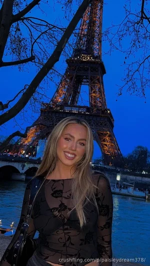 Blonde woman in sheer black floral top posing in front of Eiffel Tower at dusk.