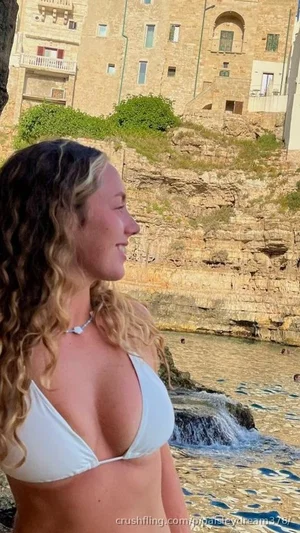 Woman with curly hair in white bikini top near rocky waterfront with buildings in background.