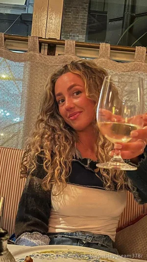 Woman with curly blonde hair holding wine glass, wearing layered top and jeans, seated at