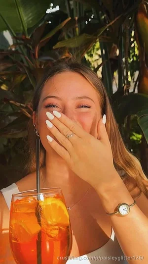 Woman with long hair covering face with hand, holding drink, wearing white top, outdoors with