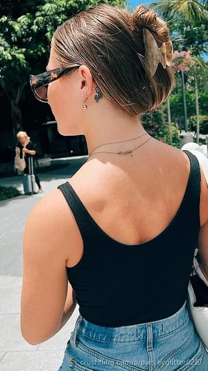 Woman in black tank top and jeans, back view, outdoors with trees and sunlight.