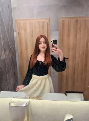 Woman with long red hair in black top and cream skirt taking mirror selfie in bathroom.