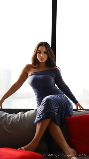 Woman in a blue one-shoulder dress sitting on a couch by a window.