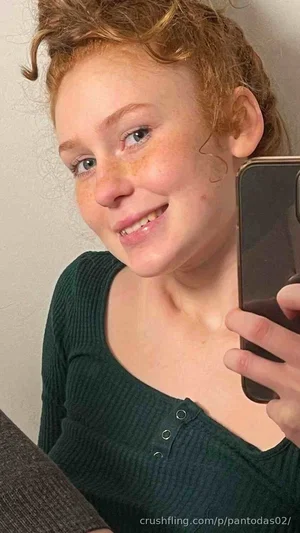 Woman with curly red hair in a green ribbed top taking a mirror selfie indoors.
