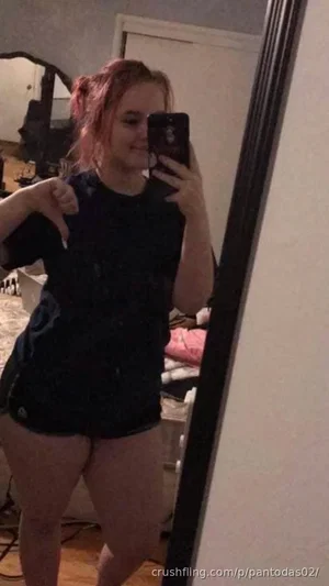 Woman with pink hair in a black t-shirt and shorts taking a mirror selfie in a bedroom.