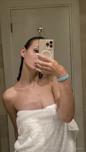 Woman with dark hair wrapped in white towel, taking mirror selfie in bathroom.
