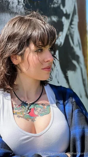 Woman with short brown hair and chest tattoo wearing white tank top and blue plaid shirt outdoors.