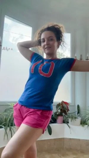 Woman in blue shirt and pink shorts posing indoors near window with plants.