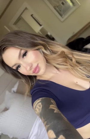 Woman with long blonde hair in blue top taking bedroom selfie.