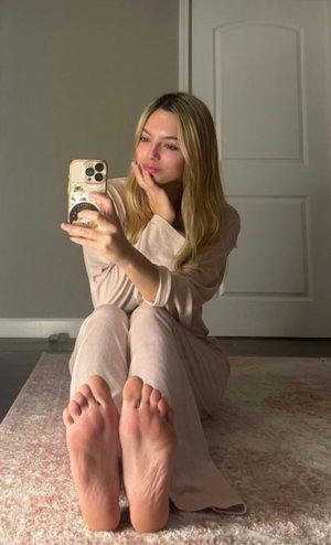 Woman with blonde hair in loose beige loungewear sits on rug, taking mirror selfie with phone.