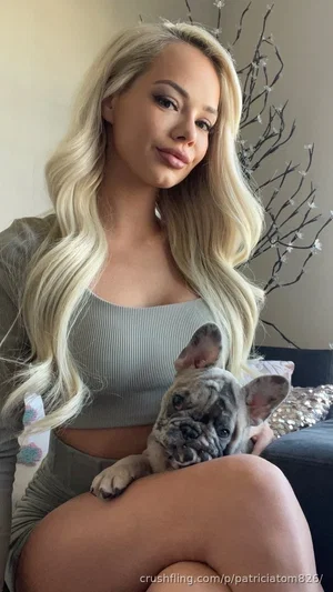 Blonde woman in gray crop top and matching pants holding a French bulldog indoors.