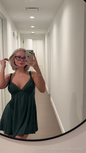 Woman with short blonde hair in green dress taking mirror selfie in hallway.