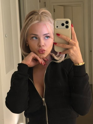 Woman with blonde hair in black zip-up jacket taking mirror selfie indoors.