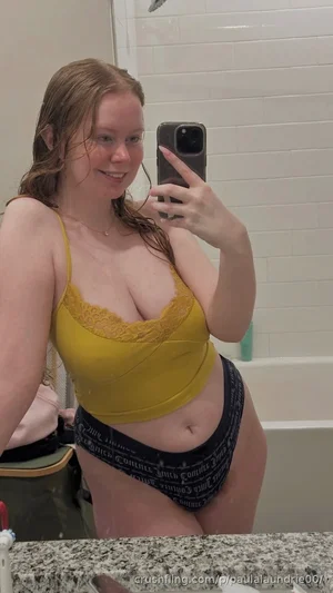 Woman in yellow lace top and patterned underwear takes mirror selfie in bathroom.