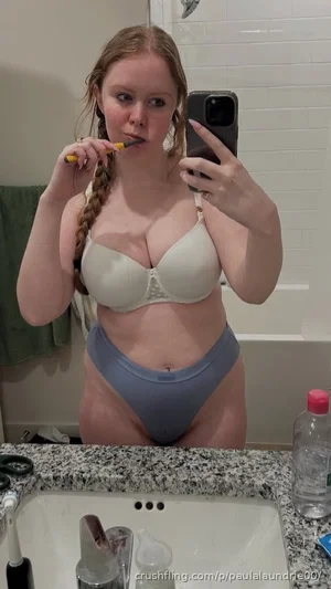Woman with braided hair in beige bra and blue underwear brushing teeth in bathroom mirror.