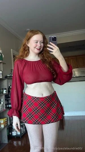 Woman with red hair in red crop top and plaid skirt, taking mirror selfie in kitchen.