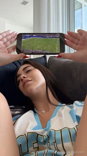 Woman in Argentina jersey lying on couch, holding phone showing soccer game.
