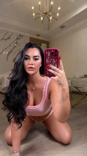 Woman with long dark hair in pink lingerie kneeling on floor in bedroom, taking mirror selfie.