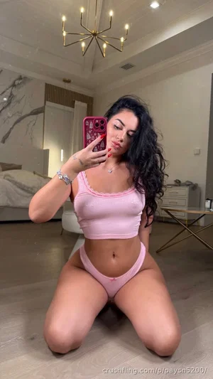 Woman with long dark hair kneeling in pink lingerie in a bedroom.