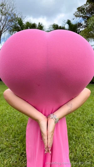 Woman in pink dress holding large pink heart outdoors in grassy area.