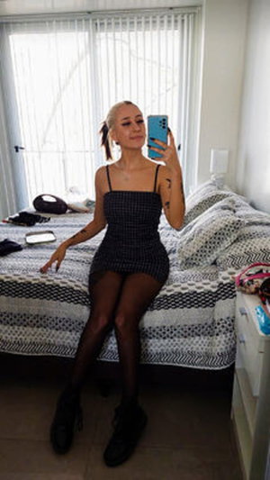 Woman in black checkered dress and sheer tights sits on bed, taking mirror selfie in bedroom.