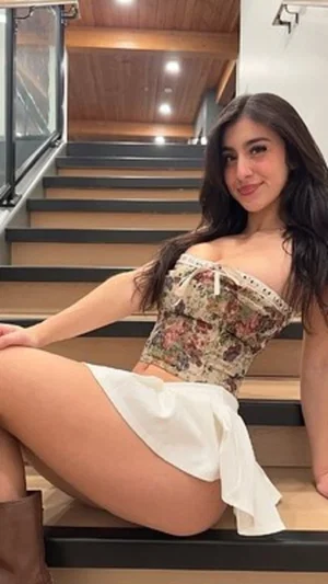 Woman with long dark hair in floral top and white skirt sitting on stairs indoors.