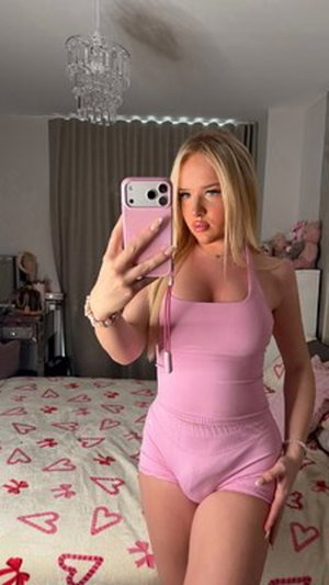 Blonde woman in pink outfit taking mirror selfie in bedroom.