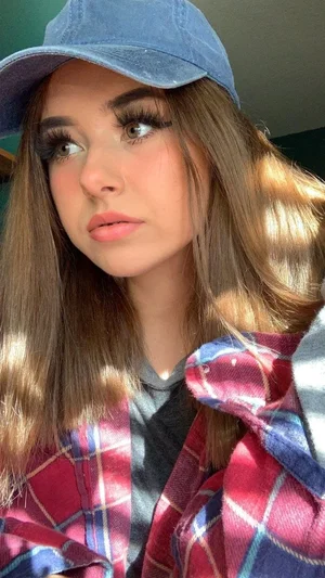 Woman with long brown hair wearing blue cap and plaid shirt, indoor selfie.