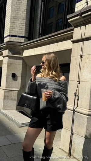 Woman in off-shoulder sweater and black shorts, holding drink, standing outside stone building.