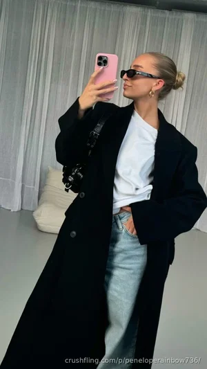 Woman in black coat, white tee, jeans, taking mirror selfie indoors.